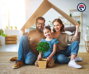 A smiling family of three, holding a cardboard cutout shaped like a house over their heads, sitting together on the floor of their home, symbolizing trust, security, and the hope of building a better future.