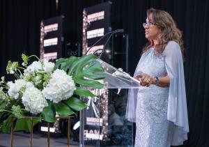 Miriam Bisnauth, Chair of the Markham Board of Trade Board of Directors, speaking at a podium adorned with white floral arrangements during the 2024 Business Excellence Awards. She is wearing a light blue lace dress with a flowing cape, addressing the aud