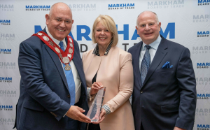  Mayor Frank Scarpitti (left), Marcia Finlayson (center), and Todd Finlayson (right) pose together, holding the Anthony Roman Award at the 2024 Business Excellence Awards. They stand in front of a backdrop displaying the Markham Board of Trade branding, s