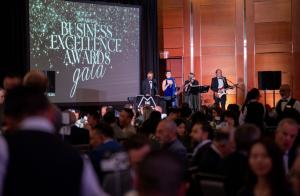 A live band performs on stage during the 34th Annual Business Excellence Awards Gala, with the event title displayed prominently on a large screen in the background. The warmly lit room is filled with elegantly dressed attendees seated at round tables, cr