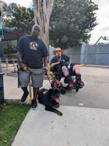 Don White, dressed in an event-specific Handicap Help, LLC’s Wheel World Consulting shirt, stands confidently holding the leash of Sir Oak Legends, a Rottweiler wearing the same event shirt. Beside them is Sgt. Jordan Teegardin, seated in a motorized whee