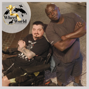 Don White, dressed in an event-specific Handicap Help, LLC’s Wheel World Consulting shirt, stands with his arms crossed beside Sgt. Jordan Teegardin, seated in a motorized wheelchair and wearing a black shirt and bright orange socks. Their confident pose