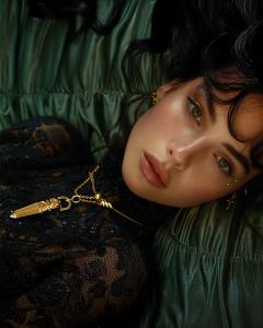 A woman with dark hair wearing the 24k gold-plated Potion Pendant as a necklace, paired with an elegant black lace outfit against a green background.