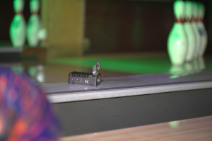 A small black edge computing device mounted on the side of a bowling lane, with bowling pins visible in the blurred background