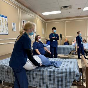 A group of CBBC Career College students in blue scrubs practicing patient care techniques in a classroom setting with medical equipment and posters visible in the background.