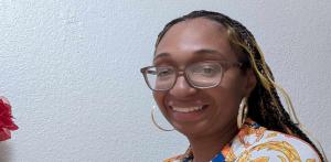 Photo of Jessica "Noonie" Noble, Editor-in-Chief of Sports and More Sports, smiling warmly while wearing glasses and a vibrant floral-patterned shirt against a neutral background. Image is featured on the webpage, "About Sports And More Sports."