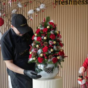 A florist designs a festive floral Christmas tree arrangement with red carnations, pinecones, gold accents, and holly berries in a glass vase