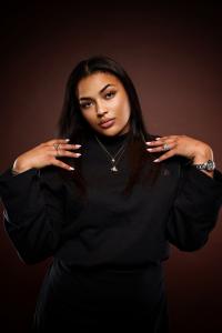 Portrait of Lia Vybz, a Fahoy Music artist, dressed in black with layered necklaces, exuding confidence and style against a warm brown backdrop.