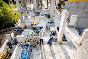 An architect reviews a blueprint while a foreman explains at a concrete construction site. In the background, partially completed structures are visible.