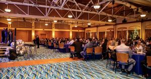 Photography of events room of SeaWorld Park in Orlando, FL. There is a stage with 3 people on the left, and rounded tables with people sitting on the right. The room is wide with wood roof and blue carpet on the floor.