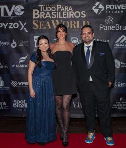 Photography of 3 people standing on a red carpet. On the left, Thays Bonini, white woman with long black hair wearing a long blue gala dress. In the middle, Thais Eliasen, white, tall woman with a short blackdress. Gero Bonini is in the right, he wears a black suit.