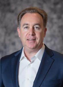 Stephen Gerard in a blue suit and white shirt stands against a gray marbled background.