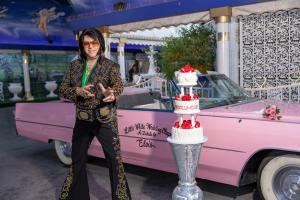 Elvis impersonator poses with three-tiered wedding cake in front of pink Cadillac at wedding drive thru with cherubs on ceiling