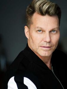 A white man in his 50's wearing a black and white jacket looking into the camera with a smirk..