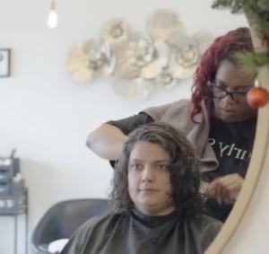 A hairstylist works on a woman's curly hair in a salon setting. A decorative wall piece and a round mirror are visible.