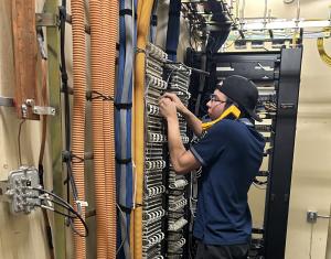 Picture showing a technician standing in a telecommunications room testing phone lines by holding a phone up to his ear.