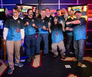 A small crowd of individuals wearing blue/black shirts who participated in the 2024 MLeS tournament, standing in an arcade