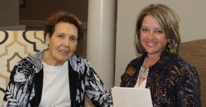 An elderly woman smiling while speaking with a care manager holding a clipboard in a cozy living room setting.