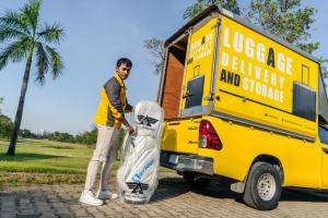 "A professional from AIRPORTELs handling a securely packed golf bag for delivery with the company’s signature yellow truck at a scenic golf course in Thailand."