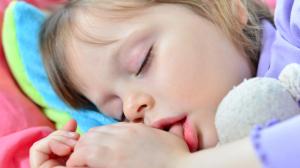 A photo of a toddler lying in bed sucking on their thumb.