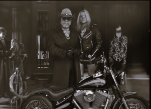 A sepia-toned image of Love Lyzardz exuding rock and rebellion. A man in a dark trench coat, cowboy hat, and gloves stands confidently beside a woman in a black leather jacket, mini skirt, and sunglasses. They are positioned in front of a sleek, chrome mo