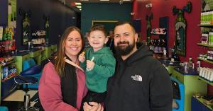 A man with a beard standing next to a boy in a green sweatshirt being held by a women in a colorful hair salon.