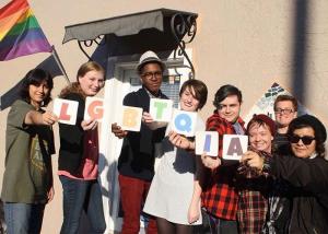 Eight diverse LGBTQ+ youth standing outside a community center, smiling and holding colorful letters that spell LGBTQIA+, with a rainbow pride flag in the background.