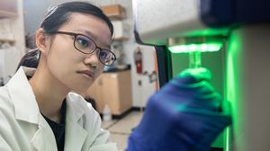 Hofstra student in lab coat and gloves holding a test tube in a machine.