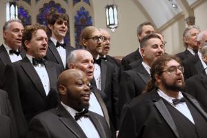 Washington Men's Camerata singing on risers in front of a church stained glass window, wearing tuxedos.