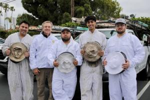 A group of five professional beekeepers from O.C. Bee Guy stand in front of their service vehicle, wearing protective suits and holding their beekeeping hats. The team is ready for a live bee removal operation.