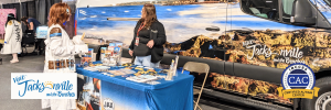 A booth promoting Jacksonville and its beaches, featuring a woman engaging with a visitor and a backdrop of a scenic van display.