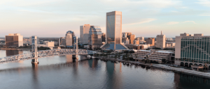 A panoramic view of Jacksonville's skyline at sunset, featuring the iconic bridge and modern skyscrapers reflecting on the river.