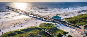 Aerial view of a long pier leading to the ocean, with sunlit waves, sandy beach, and people enjoying the seaside.