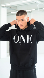 A Male Model Wearing An All Black Series I Hoodie with White Embroidered Stitching of the Word "ONE" written large across the chess in white embroidered fabric.