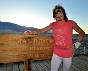 Frank McKinney standing at Badwater Basin sign in a colorful outfit with a sunset, basin landscape, and mountains in the background.