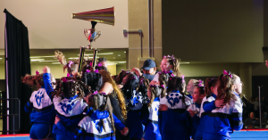 Cheer Athletics athletes celebrate on the competition floor after a victorious performance at NCA All-Star Nationals 2025. Dressed in blue and white warm-ups with the Cheer Athletics logo, the team huddles together, raising a massive trophy topped with a