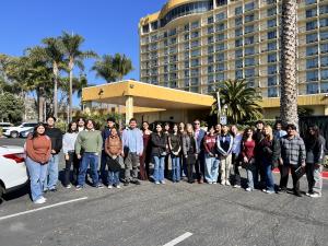 A group of 20 high school students gathered in front of the Crowne Plaza Ventura Beach, posing together with the hotel and ocean in the background.