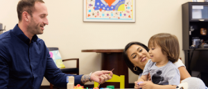 A man interacts with a young child at a table filled with toys and colorful building blocks, surrounded by a warm and engaging environment.