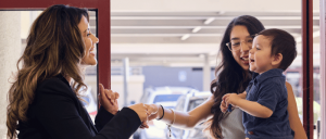 A woman and a child greet another woman at a doorway, sharing smiles and gestures amidst a parking area.