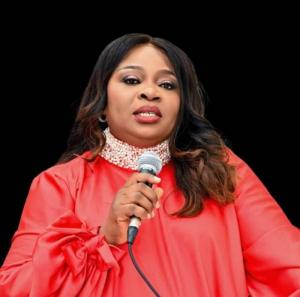 Mercy Itohan Osayi, a distinguished leader and advocate, speaks into a microphone while wearing an elegant red outfit with pearl embellishments. She is the visionary behind Women of Dignity International (WODI) and the driving force behind the Outstanding