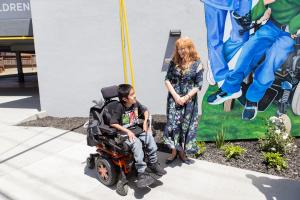 Margy Mayfield, a red haired woman in a blue floral dress, stands to the right of a young Coastal Kids Home Care patient in a wheel chair outside of the headquarters in Salinas, CA