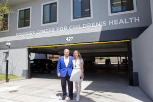 Valeta and T.J. Rodgers smile in front of a grey building and under a sign that reads "Rodgers Center for Children's Health"