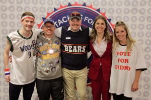 Five M4KTC volunteers posing in front of a Mustaches for Kids backdrop, dressed in creative and colorful outfits, including Skip Lindgren in a grey hoodie with campaign pins.