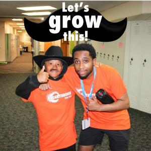 A smiling student in a cowboy hat and fake mustache poses with a mentor in matching orange Breakthrough Twin Cities shirts under a large graphic mustache with the words “Let’s Grow This!”