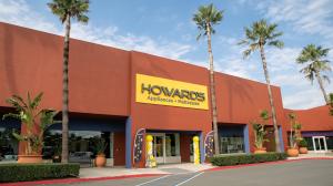 Exterior view of a Howard’s Appliances & Mattresses store with a bright yellow sign above the entrance, surrounded by tall palm trees and large potted plants. Yellow and white balloon columns flank the entrance, creating a festive atmosphere under a partl