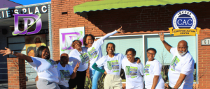 A joyful group of people in matching T-shirts celebrates outside 'JD's Place,' featuring a vibrant brick building and signage.