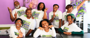 Group of eight individuals wearing matching white t-shirts with green graphics, gathered together in a cheerful pose against a purple wall.