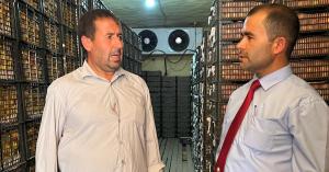 Kutbiddin Khojamurodov, wearing a blue shirt and red FINCA tie, talks with customer Nazarov Tojiddin , wearing a white buttoned shirt. They are standing inside a cold storage unit that is stacked with metal crates full of nearly ripe nectarines and peaches.