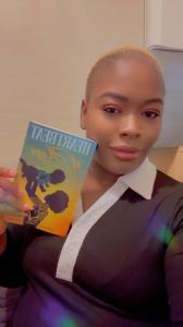 Ezinwanne is holding her book 'Heartbeat of Hope'forteh first time. She is smiling and holding the book to the camera. The cover has a silohuette of a woman with an afro holding her son uo in teh air. The heart and vessels are drawn in bright colours wher