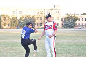 An american baseball coach in a white and red baseball uniform instructs a young Indian player in a blue DDA jersey on pitching technique on a grassy field.